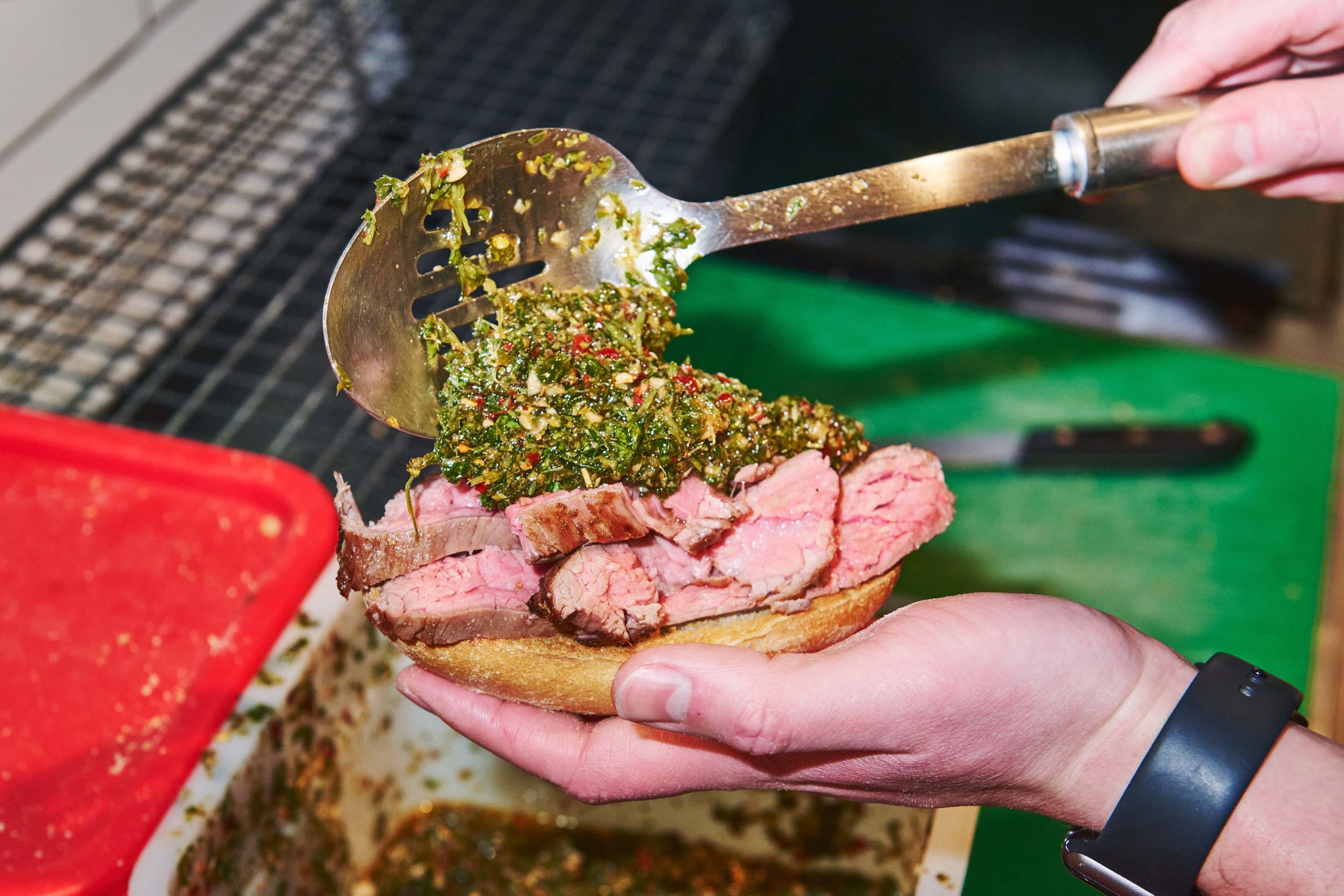 A person uses a ladle to scoop chimichurri onto a steak sandwich