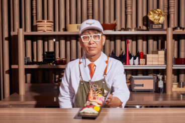 Chef Yoo-Ji Na, wearing eyeglasses and a baseball cap, presents a platter of nigiri sushi